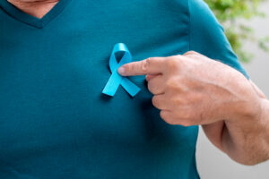 Foto em zoom de busto masculino vestido com camiseta na cor azul esverdeada, apontando para o laço, símbolo da campanha novembro azul