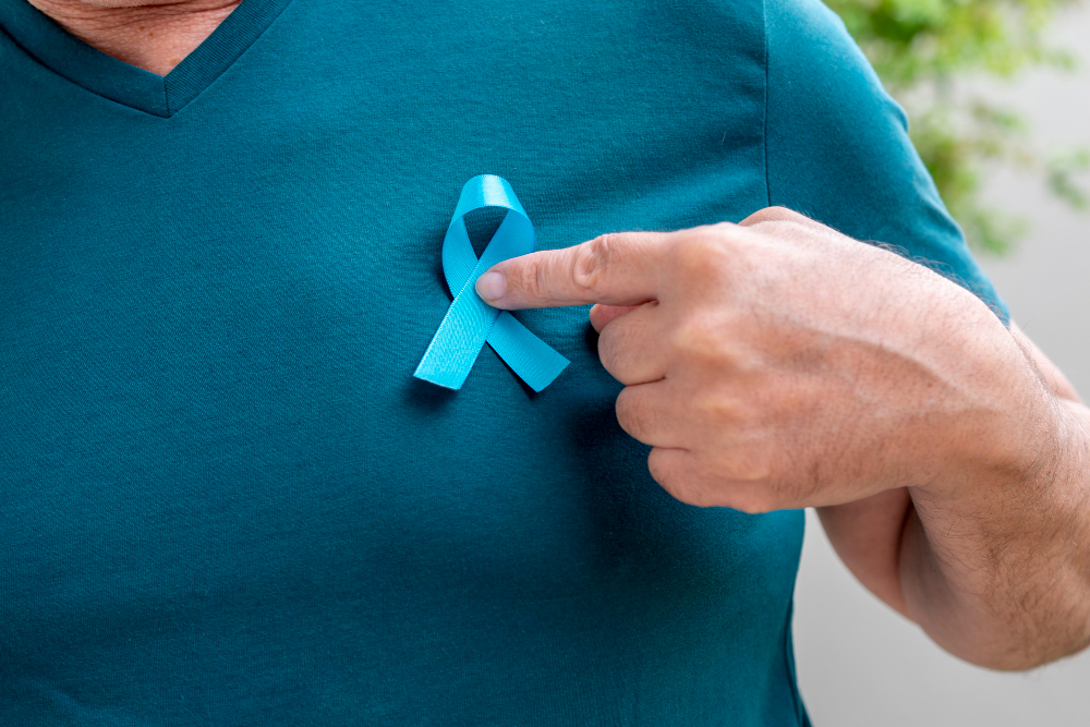 Foto em zoom de busto masculino vestido com camiseta na cor azul esverdeada, apontando para o laço, símbolo da campanha novembro azul